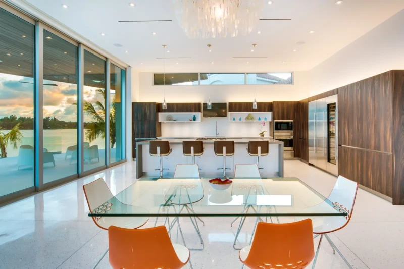 Modern luxury kitchen and dining room staging in Sarasota waterfront home with contemporary cabinetry, oversized island, glass dining table, and floor-to-ceiling windows overlooking the water
